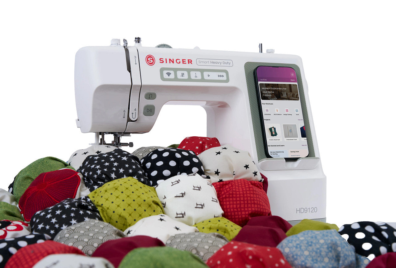A white Singer HD9120 Smart Heavy Duty sewing machine with a touchscreen display, shown on a white background with a pile of colorful fabric patches in front.