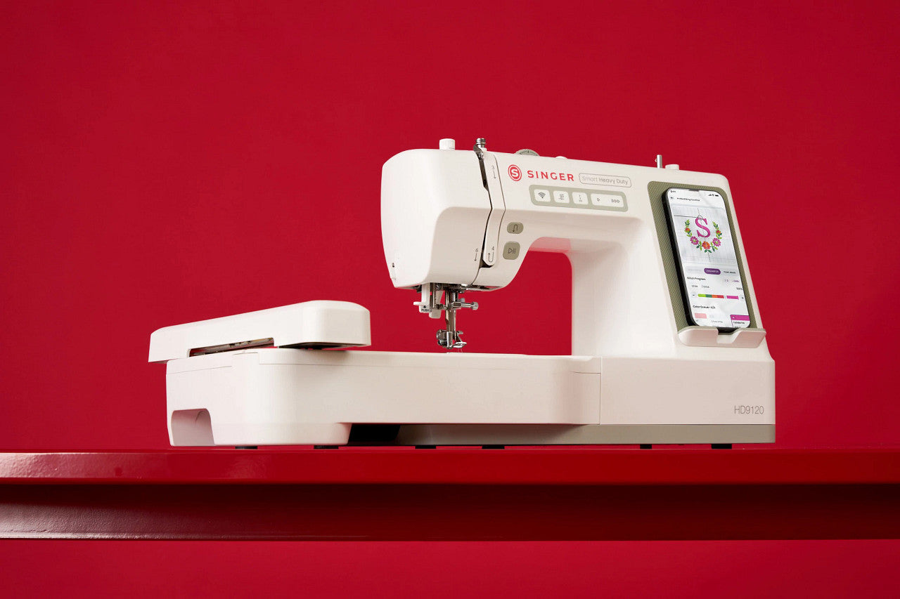 A white Singer sewing machine with an attached digital display screen, resting on a matching white quilting mat against a solid red background.