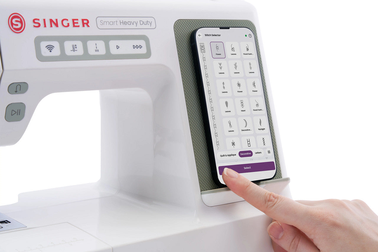 A close-up of a white Singer Smart Heavy Duty sewing machine with a smartphone app control screen mounted on the side, and a person's finger touching the screen.
