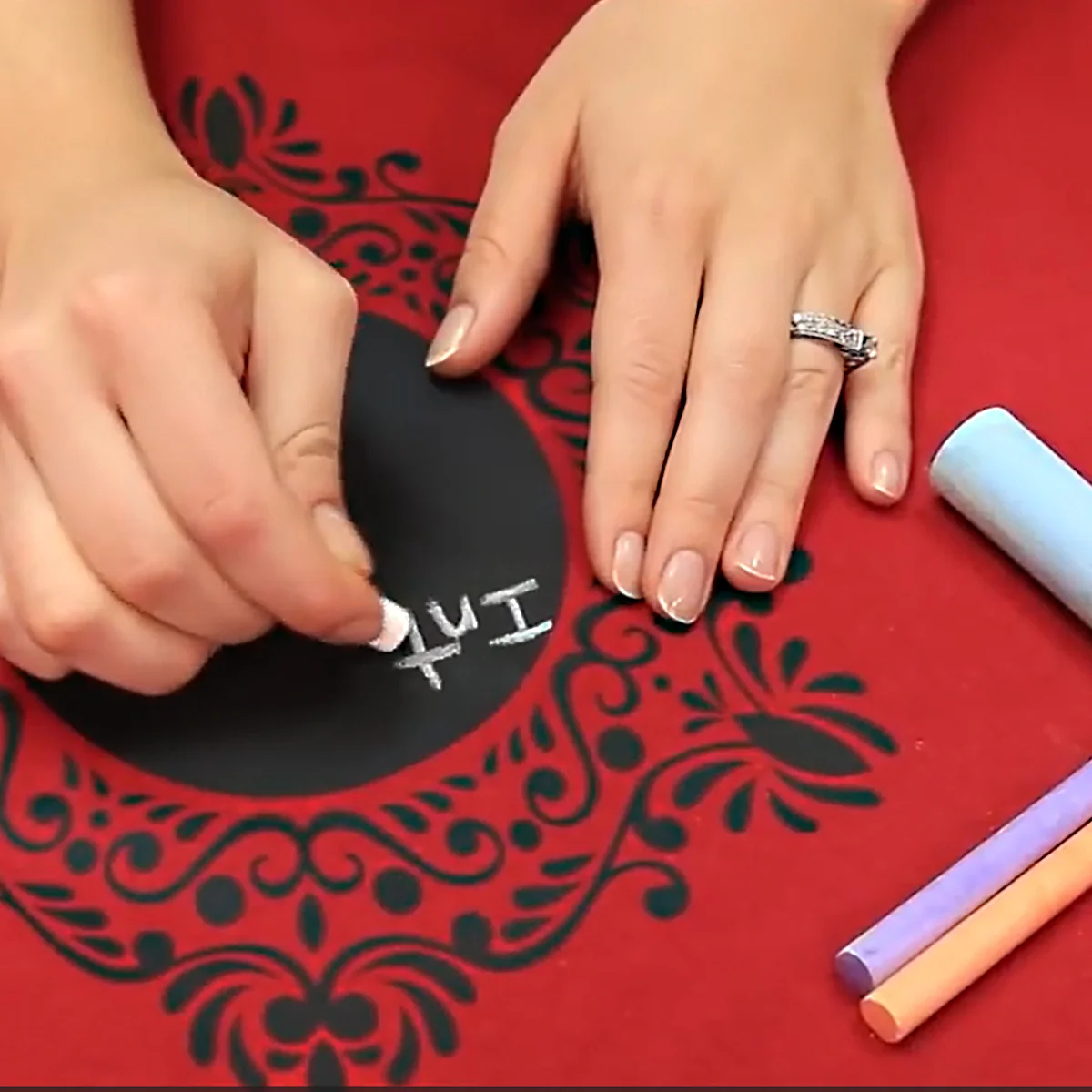Person writing on a black circular object with chalk against a red background with black patterns