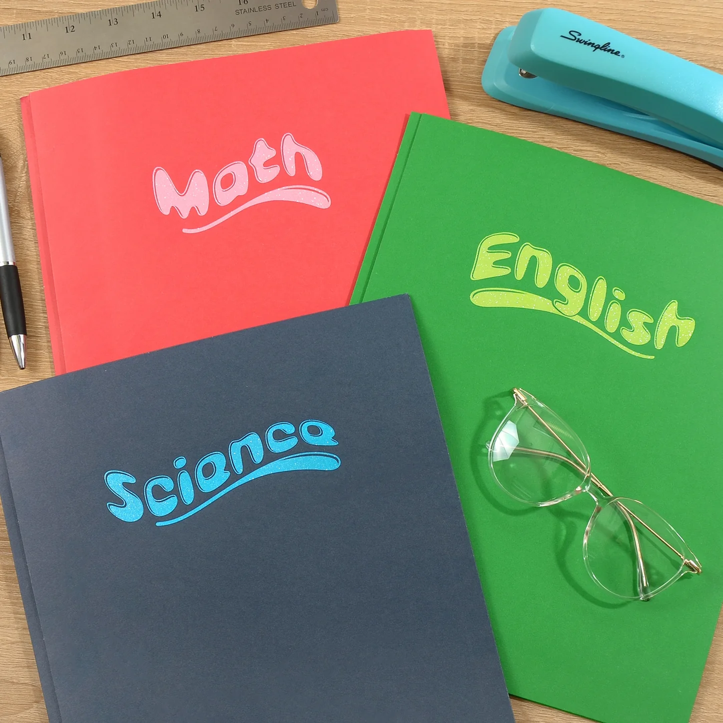 Three notebooks labeled 'Math', 'English', and 'Science' with a green notebook featuring glasses on a wooden surface.