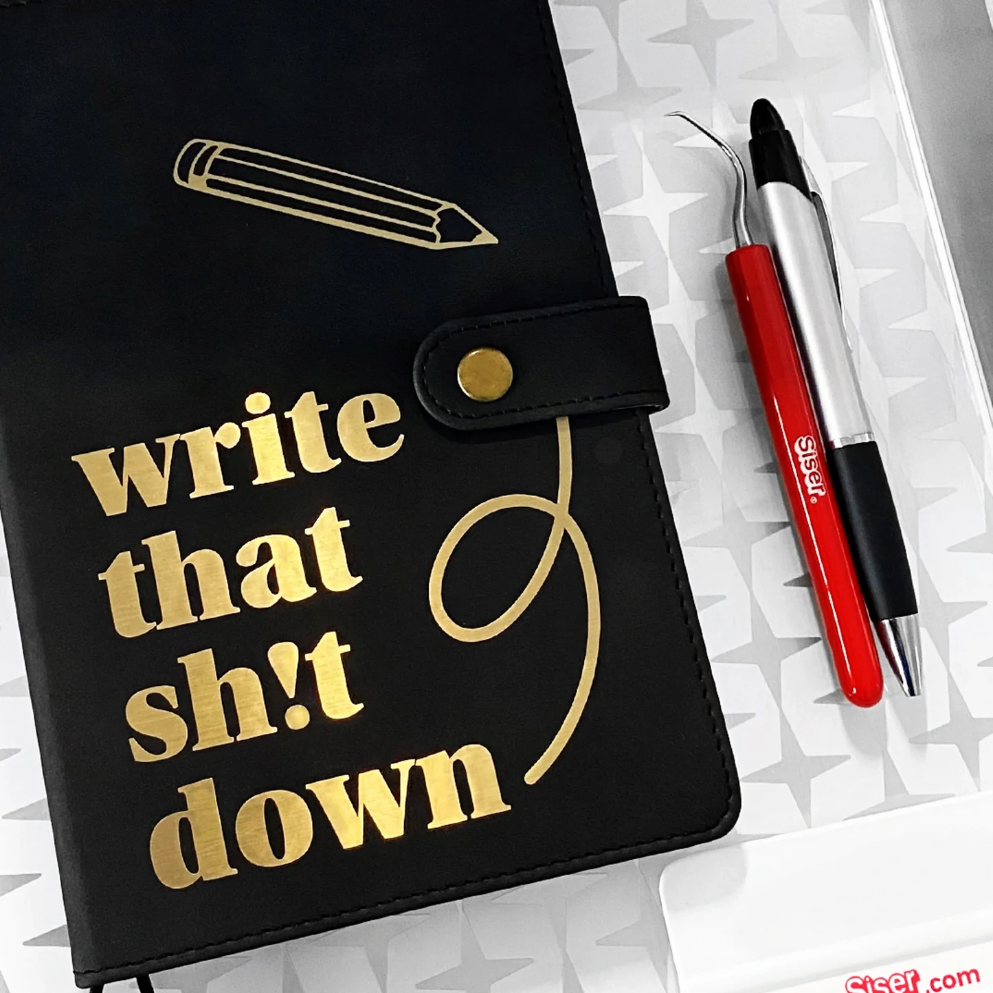 Black notebook with gold text 'write that sh*t down' and a red pen on a white background