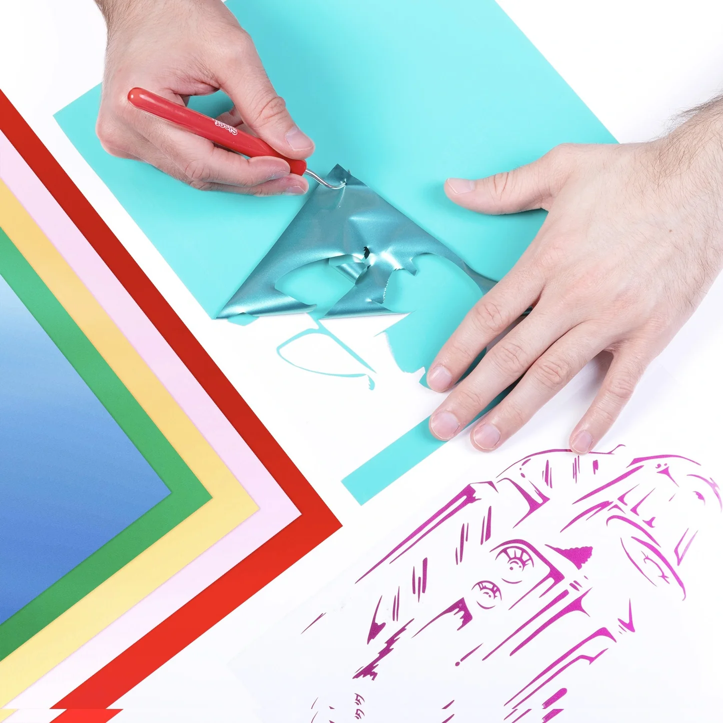 Hands using a tool to cut out a design from colored vinyl with a completed artwork in the corner.