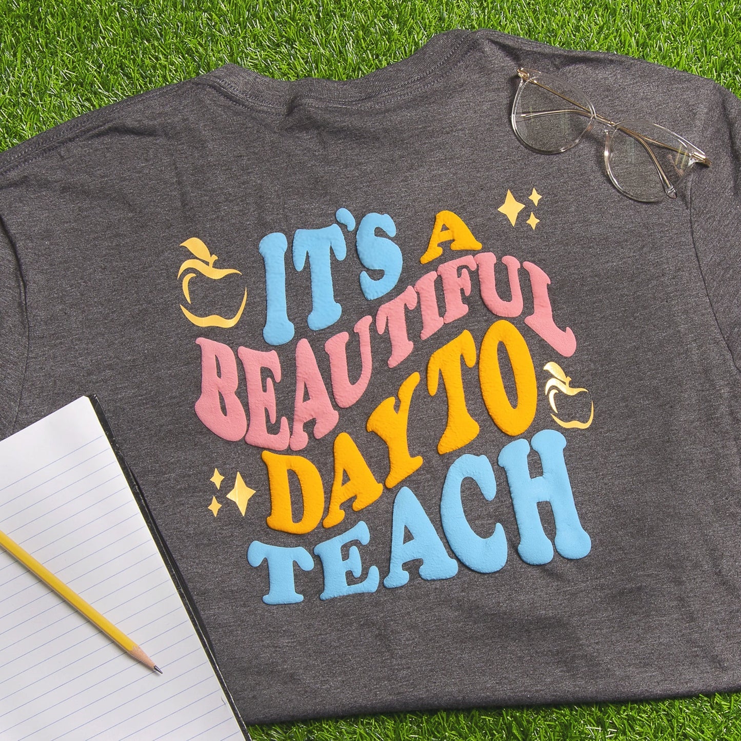 Gray t-shirt with colorful text 'It's a Beautiful Day to Teach' on grass with glasses and notebook.