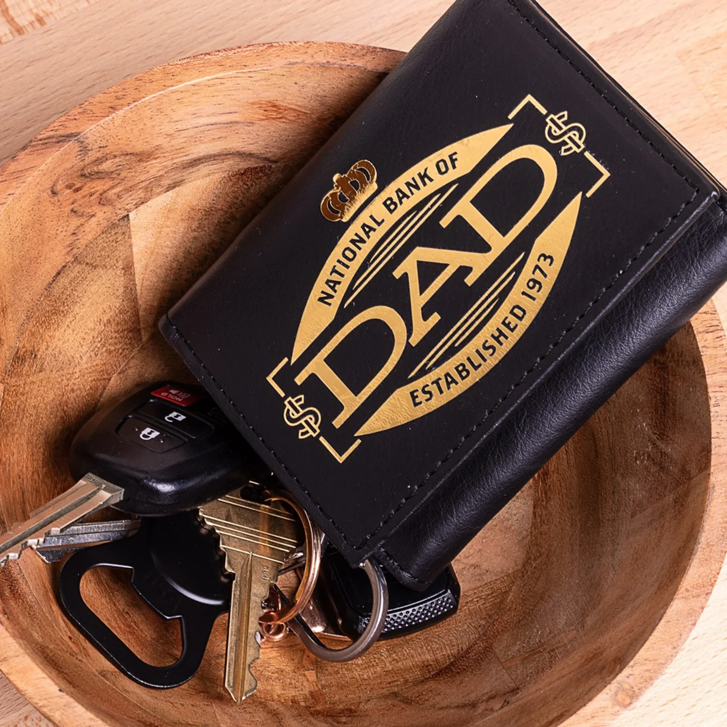 Black cardholder with 'National Bank of Dad' design on a wooden surface with keys.