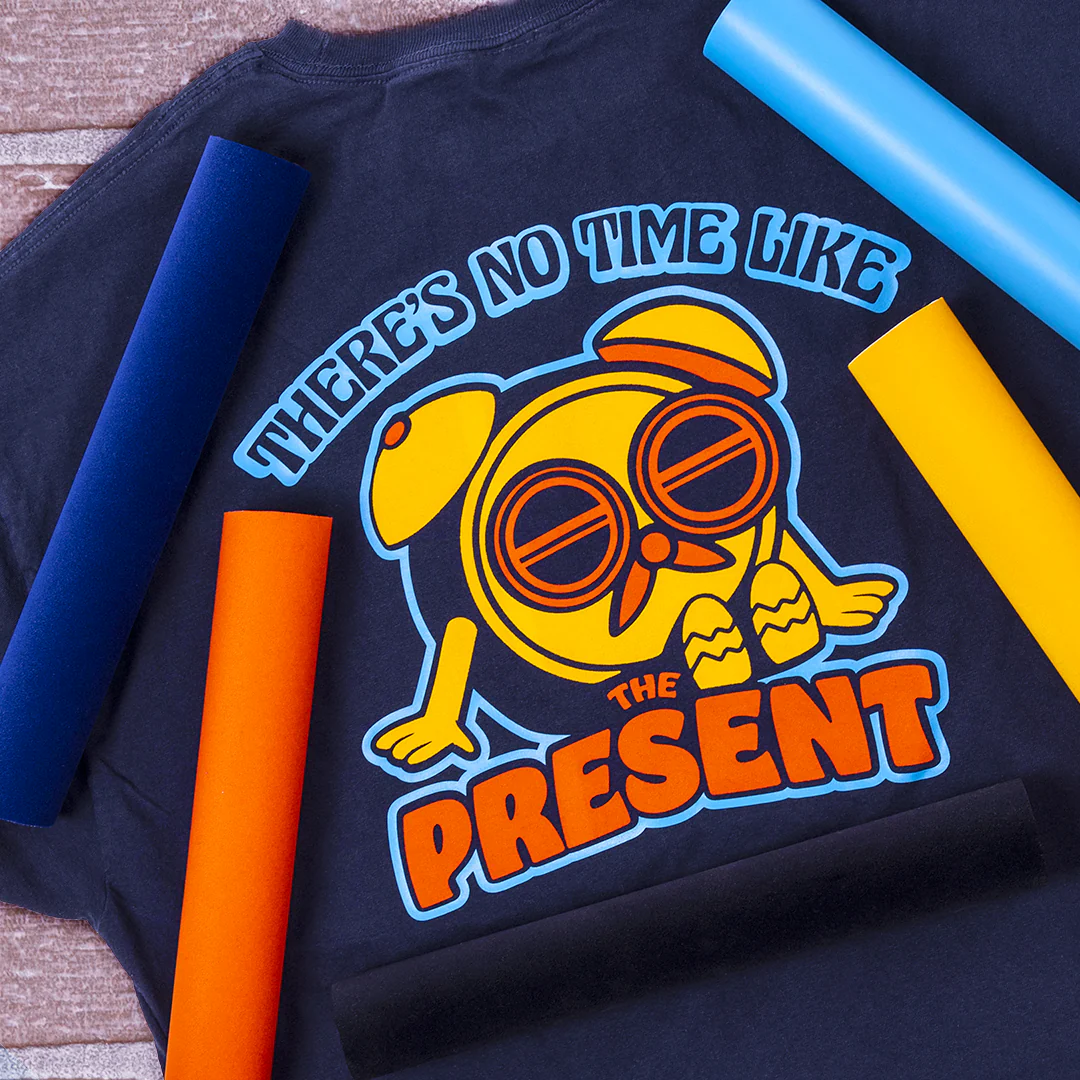 Black t-shirt with a yellow clock graphic and text 'There's no time like the present' on a wooden surface.