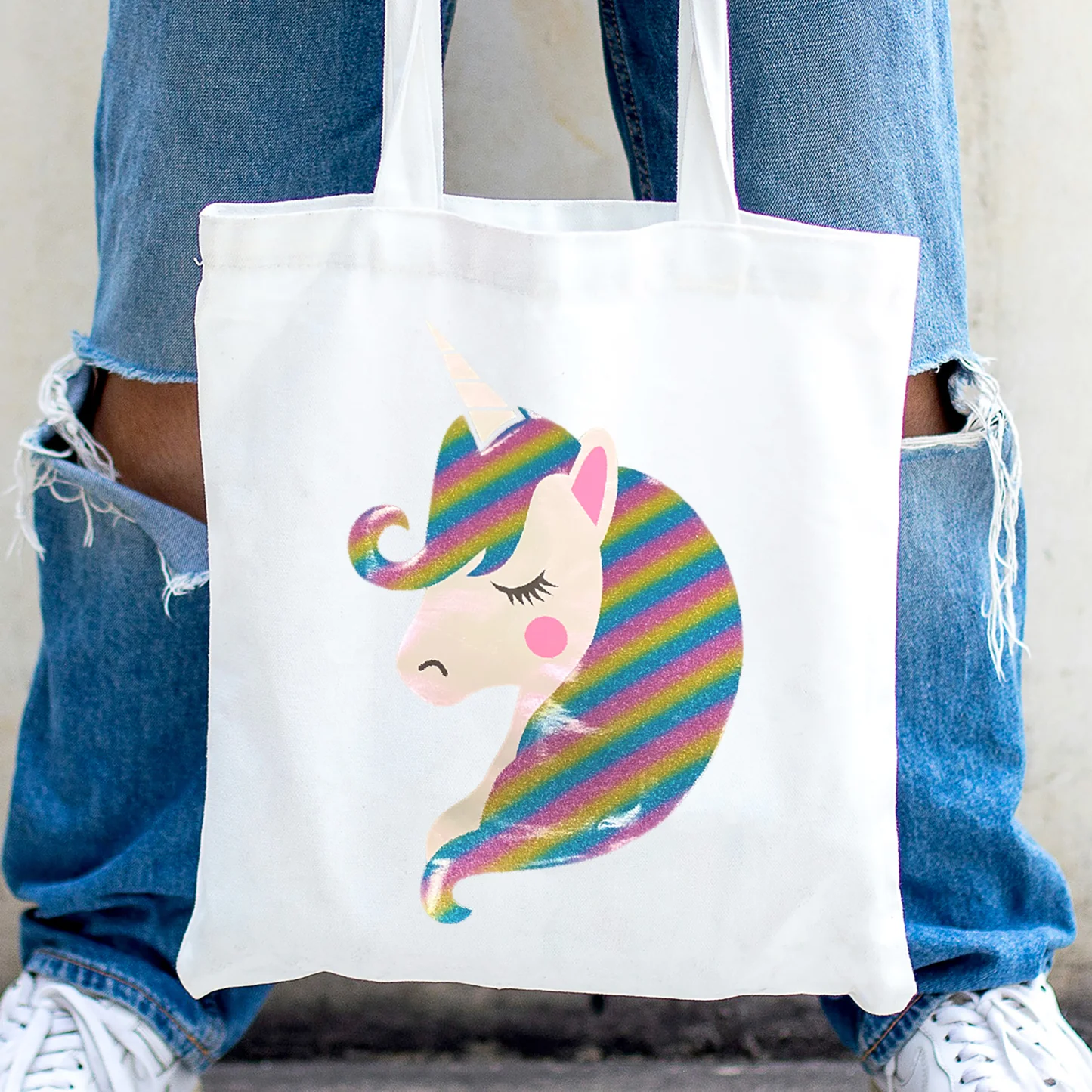 Tote bag with a colorful unicorn design held by a person wearing blue jeans and white sneakers.