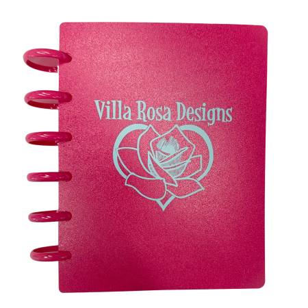 A small pink spiral-bound notebook with a matching pink wire binding, featuring the text 'Villa Rosa Designs' and a rose outline printed in light pink on the cover, shown against a plain white background.