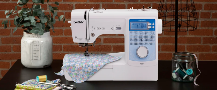 image of the Brother Innov-is NS80e six point four by four point one Computerized Sewing Machine on a table with sewing supplies and a sewing example